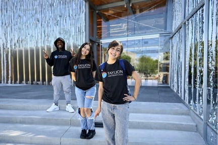 Three students wearing Taylion shirts stand smiling on outdoor steps in front of a modern school building.